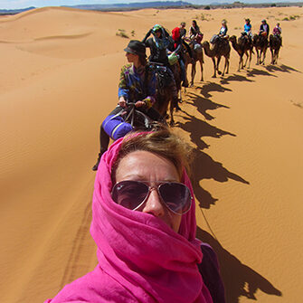 camel trek in the sahara desert, morocco, with a group of women, the first one wearing a pink scarf aournd her head and shoulders, the second one wearing a hat