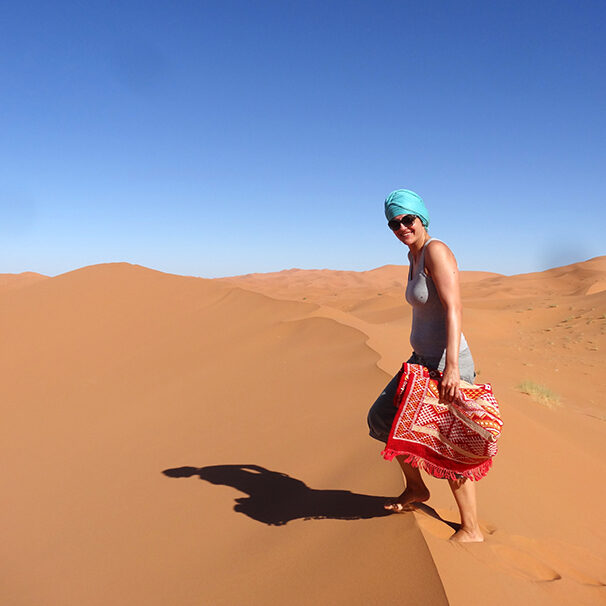 women climbing a sand dune in the sahara desert at desert u retreat, with a turquoise turban and a carpet inher hands