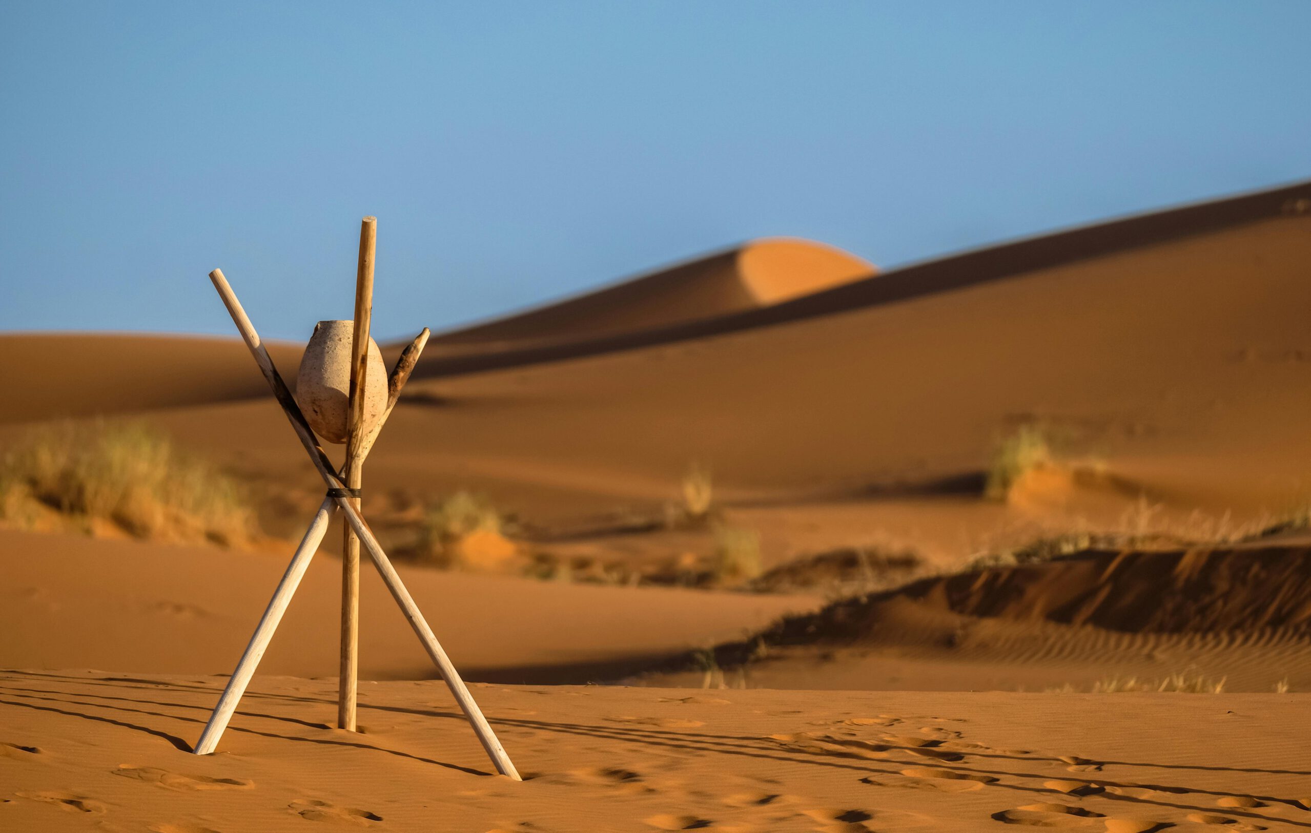 sahara desert dunes on a sunset evening, with three sticks standing up, fixed in the middle togetehr with a string, holding a ceramic pot
