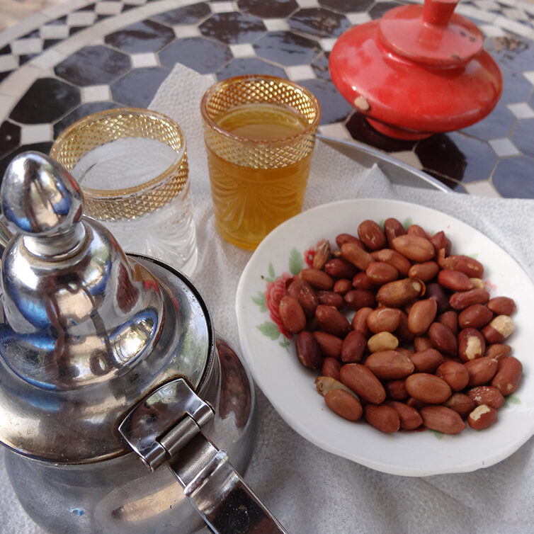 morrocan ceramic table with a metal tetiere, a plate with peanuts, two glasses with gold border and one glass filled with green tea, and a red ashtray with tap on top made of ceramics