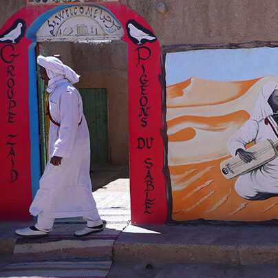 man dressed in white walking past a earth building in sahara, with a painting of gnaoua