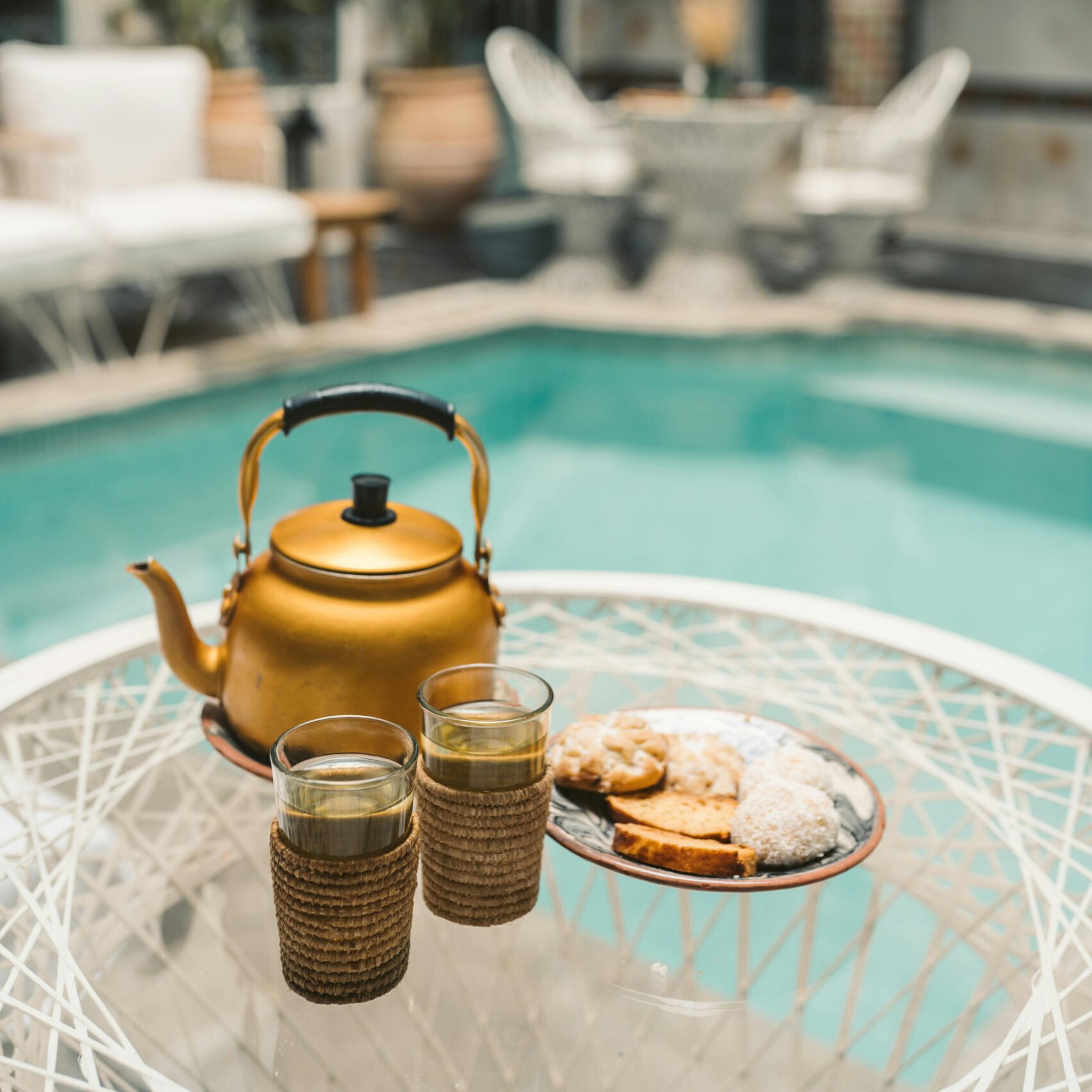 golden tea pot with two small flasses and cookies on a round glass table next to turquoise pool in traditional riad in marrakesh, morocco