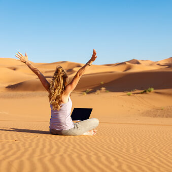 women in sahara desert, morocco, with her laptop in her knees, sitting and arms up in the air, seen from behind