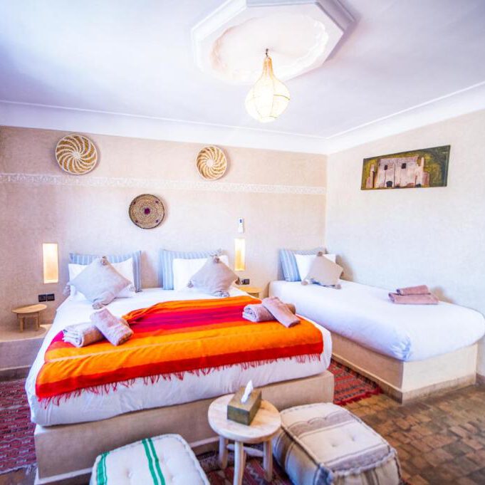 twin share room in marrakesh riad in morocco, with two beds, and one has an orange blanket on it and a small table and puffs in front of it