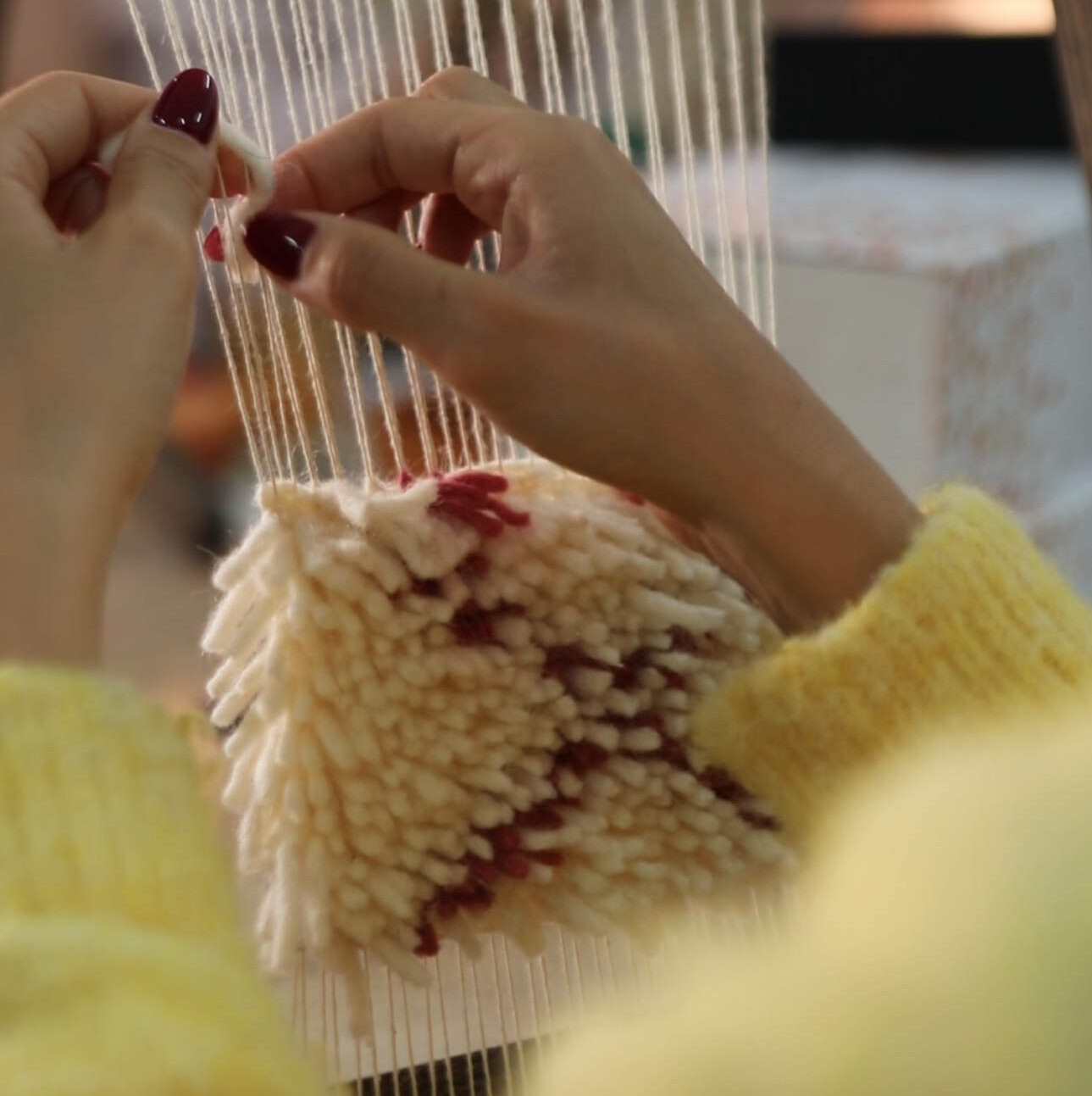 woman making a small rug / carpet on a fame, with wool, seen from behind, you only see her hands and a yellow pullower, she wears read nailpolish