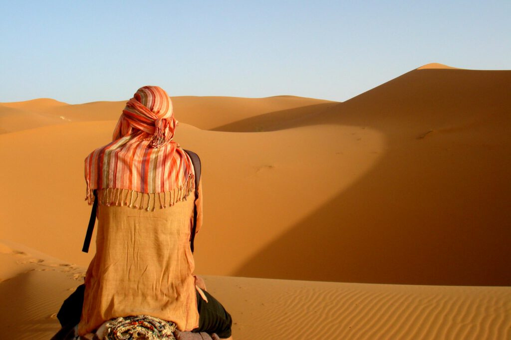 woman sitting on a dune in the sahara wearing a headscarf looking at the horizon, view from behind