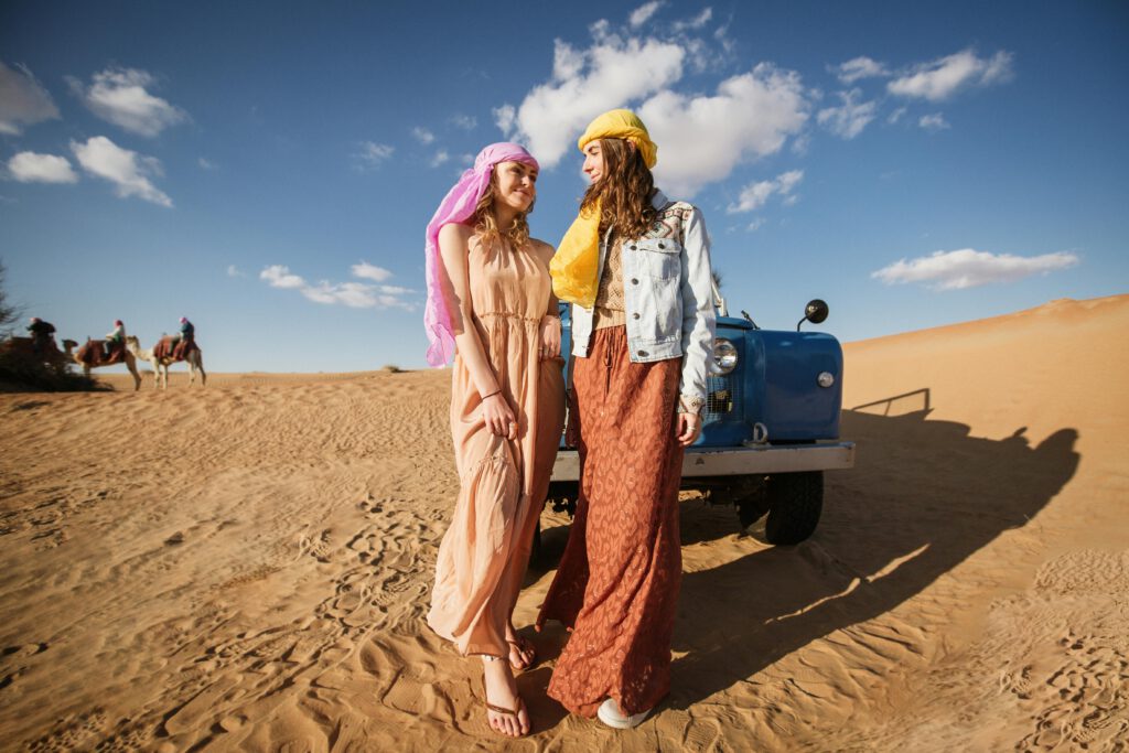 tow tourist women in the sahara desert in front of a jeep, wearing a pink and yellow headscarf and long dresses