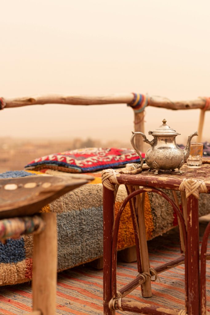 ancient arabic metal tea pot on wodden table ith red pillow on a seat in the background in the mountains or desert area