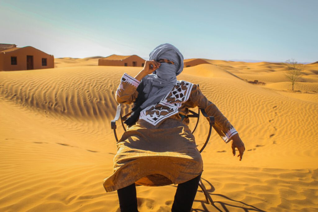 berber guy wearing a grey headscarf covering his face, sitting on a chair in the desert, wearing a brown jilaba over jeans