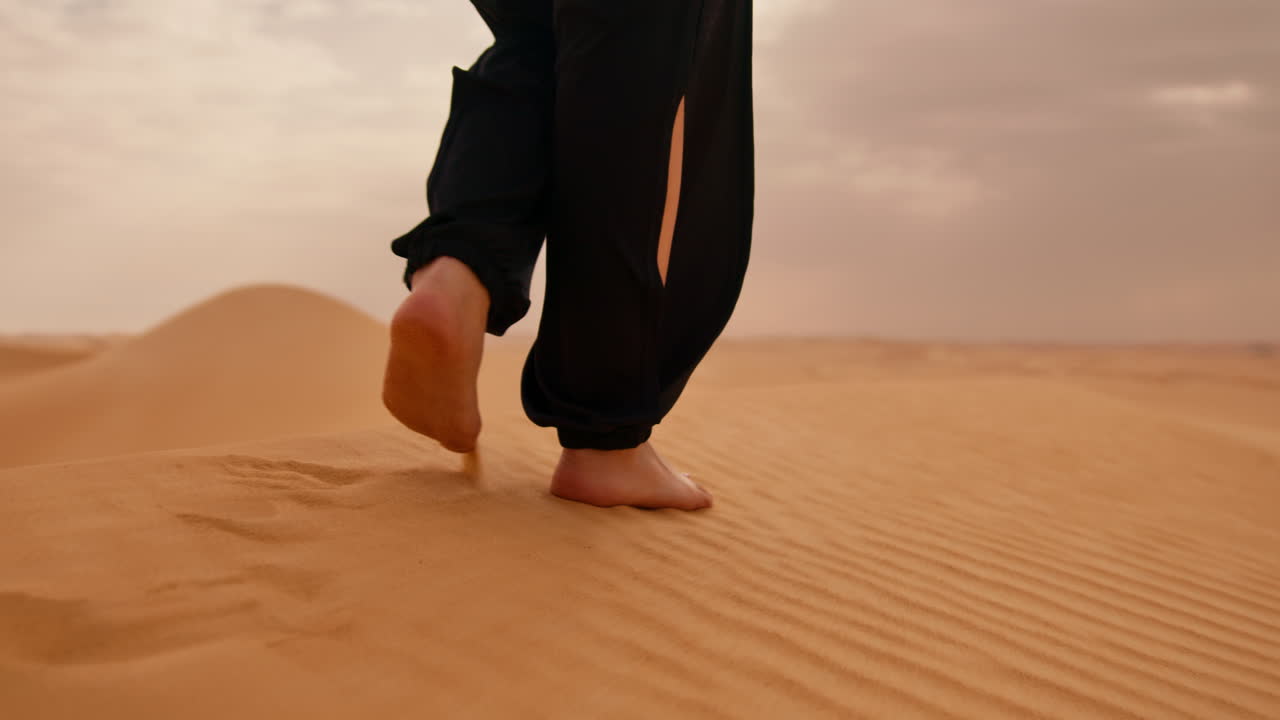 person walking barefoot in the sahara desert, with black pants, you see the feet and the chins / calve muscles